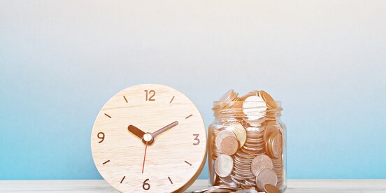 Ein Glas mit Münzen neben einer Holz-Uhr auf einem Tisch – das Bild steht symbolisch für die Betriebsratsvergütung und die Verbindung von Zeitaufwand und finanzieller Bewertung.