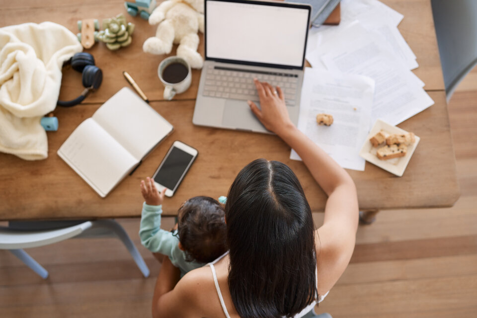 Eine Mutter arbeitet im Homeoffice am Laptop, während sie ihr Kind auf dem Schoß hält – ein Sinnbild für die Herausforderung zwischen Care-Arbeit und beruflicher Teilhabe.