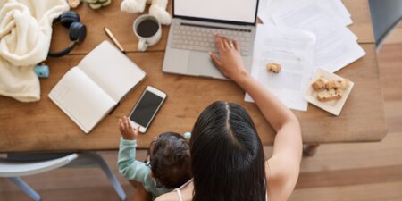Eine Mutter arbeitet im Homeoffice am Laptop, während sie ihr Kind auf dem Schoß hält – ein Sinnbild für die Herausforderung zwischen Care-Arbeit und beruflicher Teilhabe.