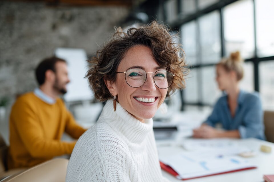 Frau mit weißem Pullover und Brille sitzt im Meeting mit zwei Kollegen