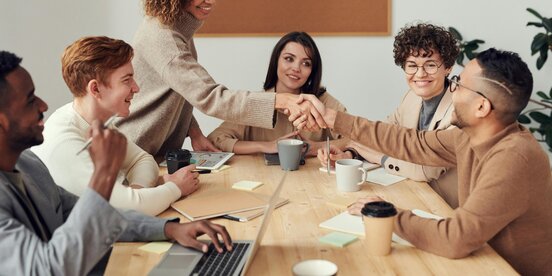 Ein fröhliches Team in einem Meetingraum, zwei Personen geben sich zur Begrüßung die Hand – das Bild symbolisiert gelungene Zusammenarbeit und erfolgreiche Klausurtagung im Betriebsrat.