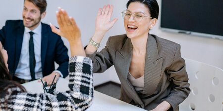 Zwei Frauen geben sich im Büro ein High Five, während ein Kollege im Hintergrund lächelt – das Bild steht für erfolgreiche Zusammenarbeit und gelebte Inklusion im Arbeitsumfeld. Zwei Frauen geben sich im Büro ein High Five, während ein Kollege im Hintergrund lächelt – das Bild steht für erfolgreiche Zusammenarbeit und gelebte Inklusion im Arbeitsumfeld.