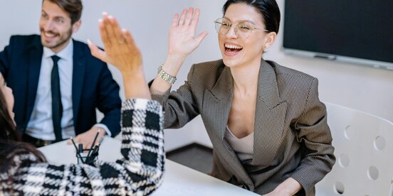 Zwei Frauen geben sich im Büro ein High Five, während ein Kollege im Hintergrund lächelt – das Bild steht für erfolgreiche Zusammenarbeit und gelebte Inklusion im Arbeitsumfeld. Zwei Frauen geben sich im Büro ein High Five, während ein Kollege im Hintergrund lächelt – das Bild steht für erfolgreiche Zusammenarbeit und gelebte Inklusion im Arbeitsumfeld.
