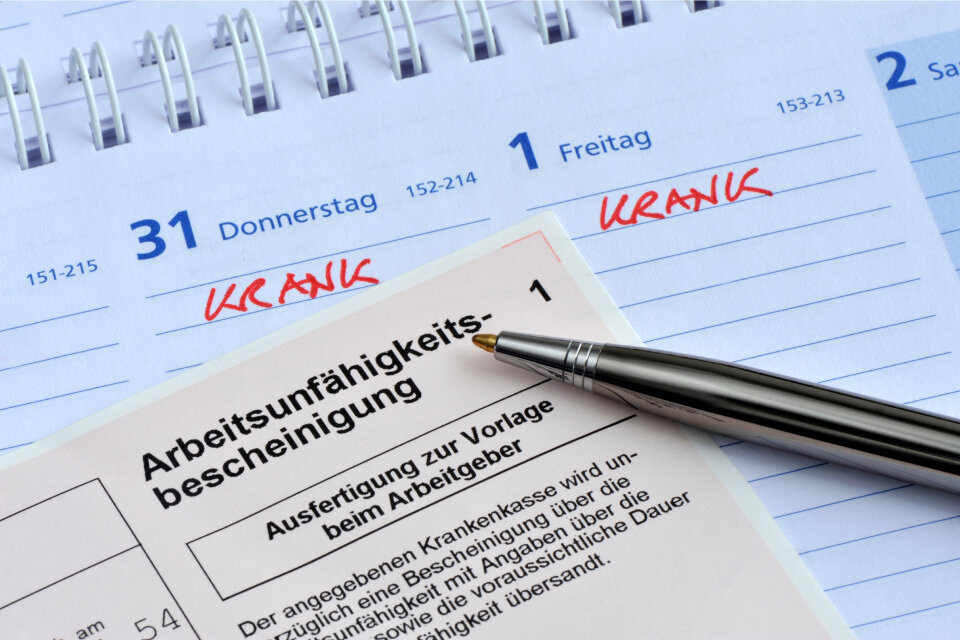 Arbeitsunfähigkeitsbescheinigung mit Kugelschreiber auf einem Kalender, in dem mehrere Tage als „krank“ markiert sind – Symbolbild für Entgeltfortzahlung im Krankheitsfall. Arbeitsunfähigkeitsbescheinigung mit Kugelschreiber auf einem Kalender, in dem mehrere Tage als „krank“ markiert sind – Symbolbild für Entgeltfortzahlung im Krankheitsfall.