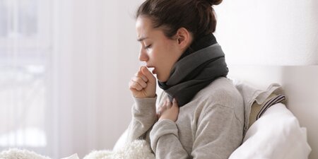 Erkrankte Frau mit Schal sitzt hustend im Bett – Symbolbild für Krankheit, Arbeitsunfähigkeit und das Recht auf Privatsphäre gegenüber Arbeitgeberbesuchen. Erkrankte Frau mit Schal sitzt hustend im Bett – Symbolbild für Krankheit, Arbeitsunfähigkeit und das Recht auf Privatsphäre gegenüber Arbeitgeberbesuchen.