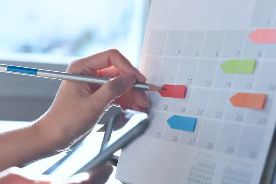 Hand markiert ein Datum auf einem Kalender mit bunten Markierungen – Symbol für den Wahlkalender der Betriebsratswahl. Hand markiert ein Datum auf einem Kalender mit bunten Markierungen – Symbol für den Wahlkalender der Betriebsratswahl.