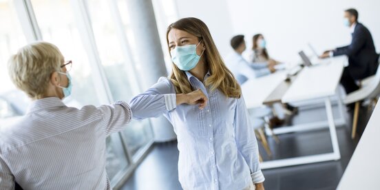 Zwei Frauen tragen eine Corona Maske und geben sich einen Ellbogen-Check