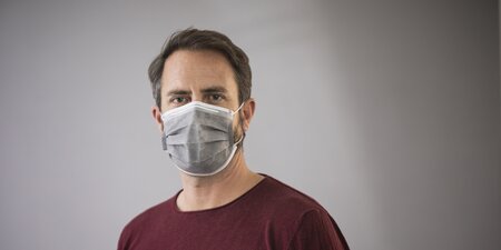 Ein Mann steht vor einer grauen Wand und trägt eine Corona Maske Ein Mann steht vor einer grauen Wand und trägt eine Corona Maske