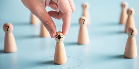 Hand wählt eine Spielfigur mit einer Glühbirne aus einer Gruppe mit Fragezeichen – Symbol für das Gewinnen von Kandidaten bei der Betriebsratswahl.