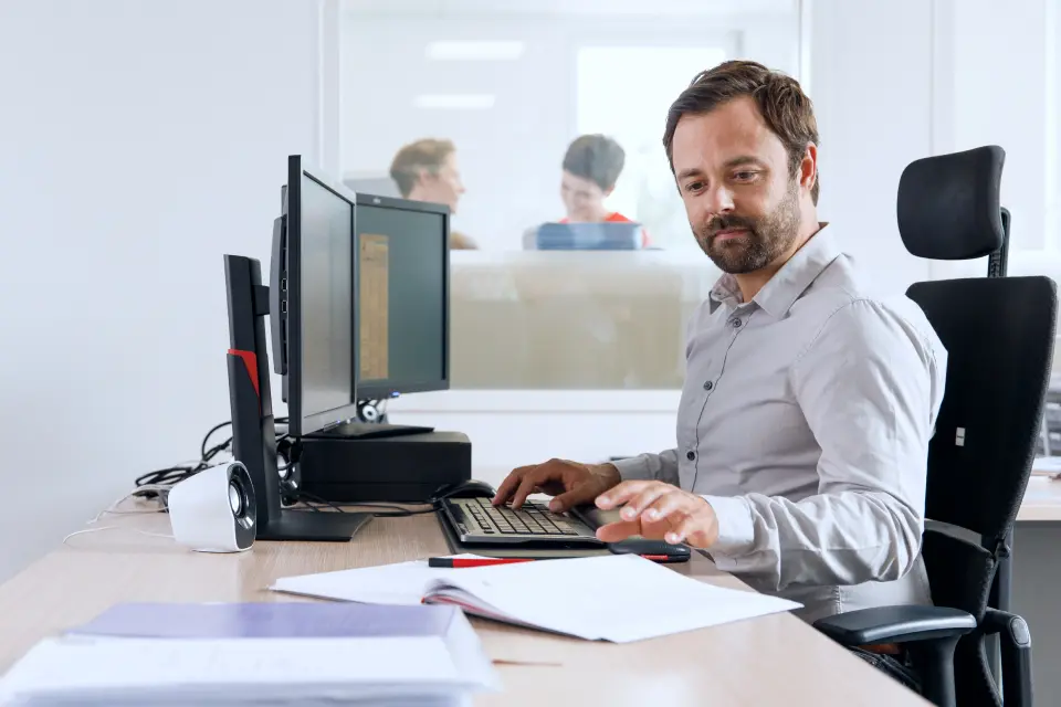 Mann arbeitet am Computer mit Dokumenten auf dem Schreibtisch – Symbol für die Berechnung der Betriebsratsgröße. Mann arbeitet am Computer mit Dokumenten auf dem Schreibtisch – Symbol für die Berechnung der Betriebsratsgröße.
