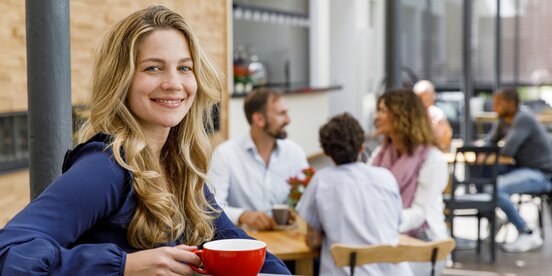 Eine Frau sitzt an einem Tresen mit einer Tasse. Im Hintergrund sind Menschen an Tischen mit Tassen zu sehen.